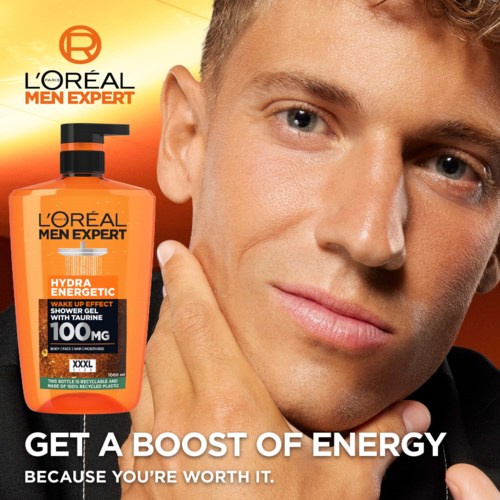 A young man with short hair and clear skin, smiling gently, next to a bright orange bottle of men's skincare product.