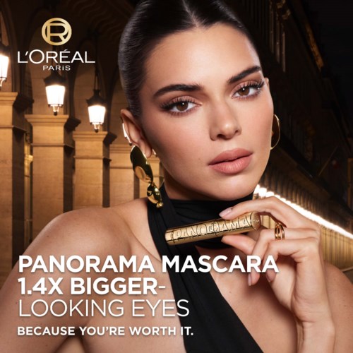 A woman with bold makeup and jewelry holds a mascara tube in a city scene. The text promotes L'Oréal mascara for bigger eyes.