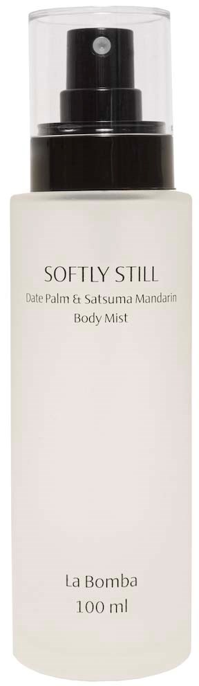 Hvit flaske med Softly Still kroppsspray med svart spraymunnstykke, merket med duftnoter og merkenavn.