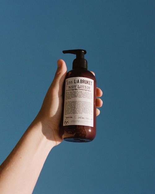 Eine Hand hält eine braune Flasche mit Körperlotion und Pumpe vor einem einfachen blauen Hintergrund.