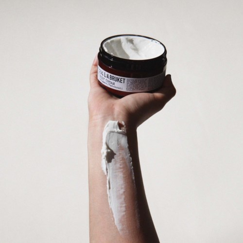 A hand holding a jar of creamy white substance, with some cream spread along the forearm. Plain background.