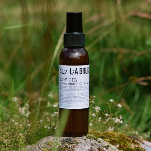 Een kleine bruine sprayfles met label 'L:A BRUK' met lichaamsolie, buiten op een rots met gras in de achtergrond.