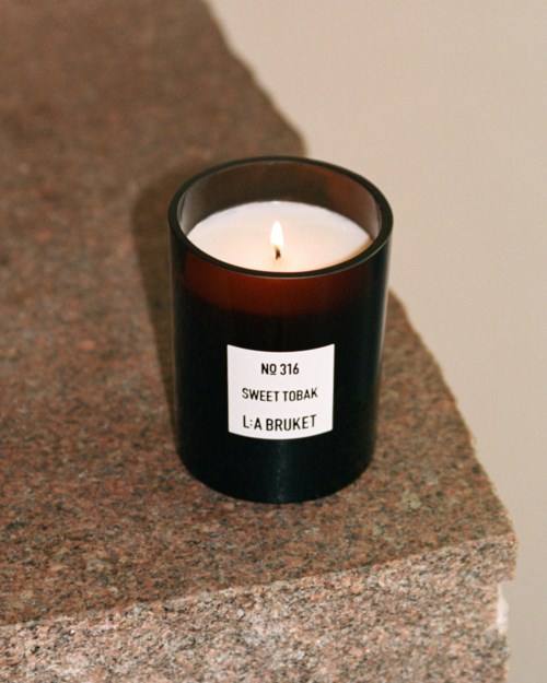 Een zwart kaarsenpotje met een brandende witte kaars op een stenen oppervlak. Label zegt "NO 316 SWEET TOBAK L:A BRUKET."