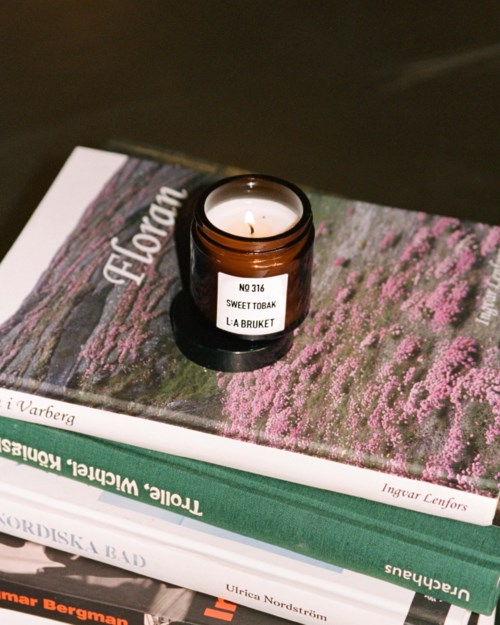 Een kleine bruine kaarsenpot met een brandende kaars op gestapelde boeken, waaronder een getiteld 'Torleif Rindal' en 'Ursula Nordström.'