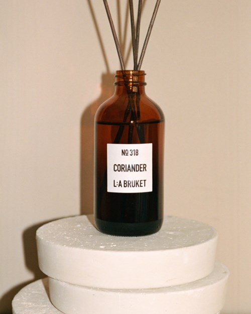 Eine braune Flasche mit Stäbchen auf einem weißen runden Sockel, beschriftet 'Koriander La Bruket', vor beigem Hintergrund.