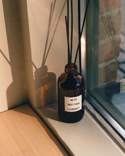 Bruine fles met zwarte stokjes op vensterbank, label zegt "Sweet Tobak."