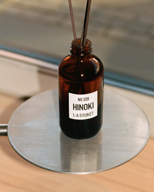 Eine braune Flasche mit der Aufschrift 'HINOKI LA BRUKET' steht auf einer runden Metallfläche in der Nähe eines Fensters.