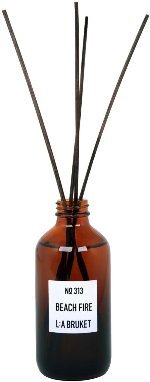 Een bruine glazen fles met een label en vijf dunne stokjes die eruit steken. Het label zegt "BEACH FIRE" en "L:A BRUKET."