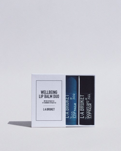 Eine weiße Box enthält zwei Lippenbalsamstifte, eines blau und eines schwarz, mit Labels 'Wellbeing Lip Balm Duo' und 'La Bruket.'