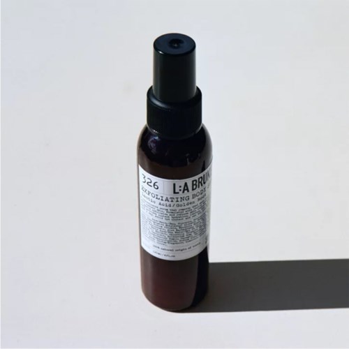 Eine kleine braune Sprühflasche mit schwarzem Deckel und weißem Etikett, auf einer weißen Oberfläche mit Schatten.
