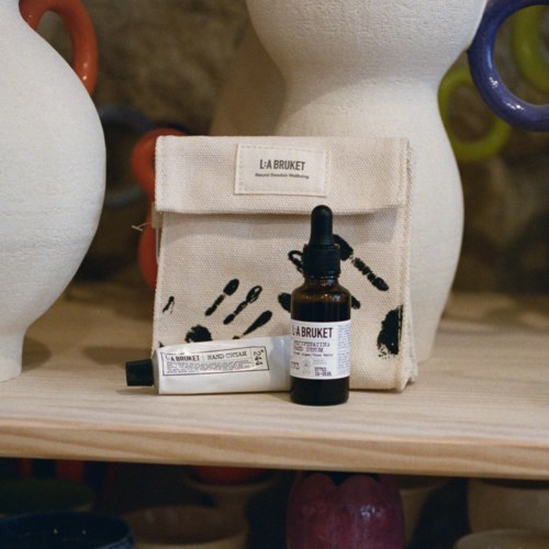 Eine kleine Tasche und eine Flasche Handdesinfektionsmittel von La Bruket stehen auf einer Holzeplatte. Die Tasche hat schwarze Schrift und Bilder.