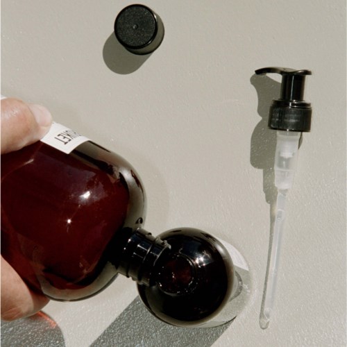 Eine Person hält eine braune Flasche in der Nähe einer Wand mit einem Seifenspender und einem kleinen schwarzen Deckel.