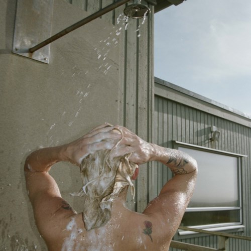 Person wäscht Haare im Freien mit Wasser aus einer Dusche neben einem grauen Gebäude.