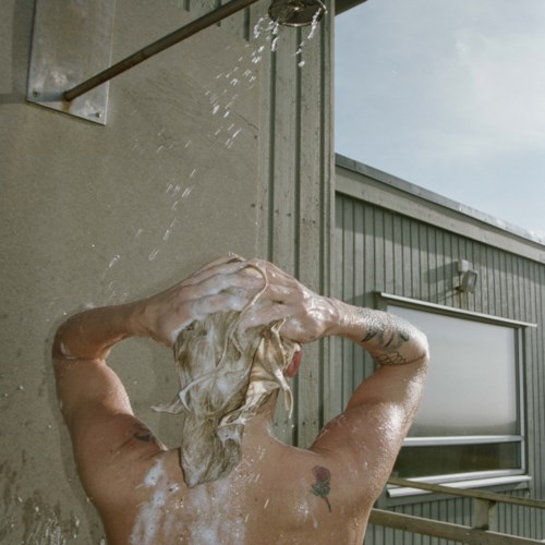 Eine Person duscht draußen und wäscht sich die Haare unter einem Wasserstrahl an der Wand.
