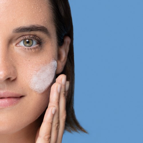 A woman applies facial cleanser to her cheek, with a soap lather on her face, against a blue background.