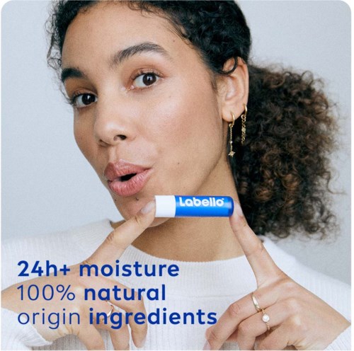Een vrouw houdt een Labello lipbalsem vast, wijst ernaar, met tekst over meer dan 24 uur vocht en natuurlijke ingrediënten.