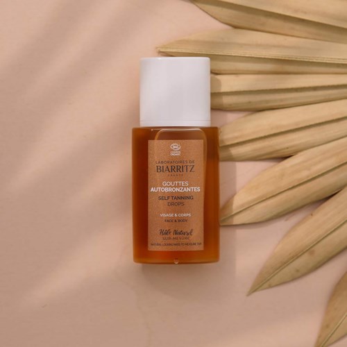 En liten orangebrun flaska med vit topp, märkt för self-tanning drops av Biarritz, omgivet av ljusa blad.