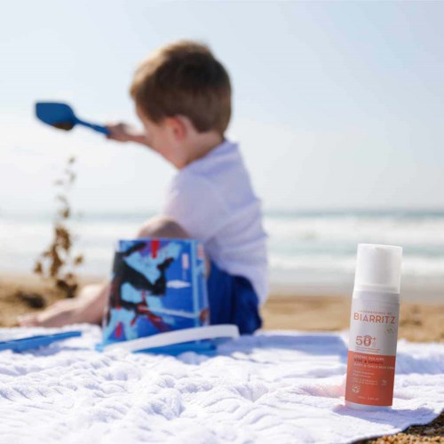 A child on the beach playing in the sand with a shovel, beside a bottle of SPF sunscreen. The sea is in the background.