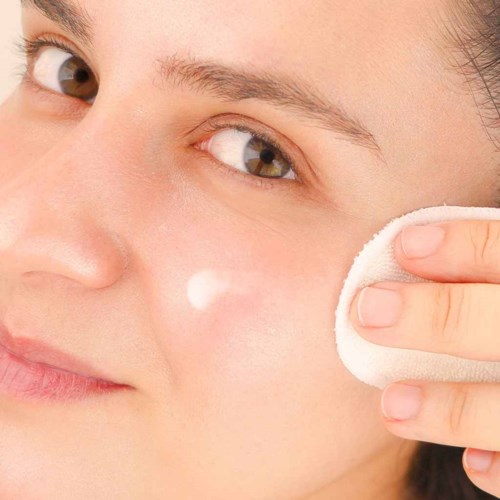 A person applies cream to her face using a cotton pad, smiling gently. Close-up of skin and eye details.