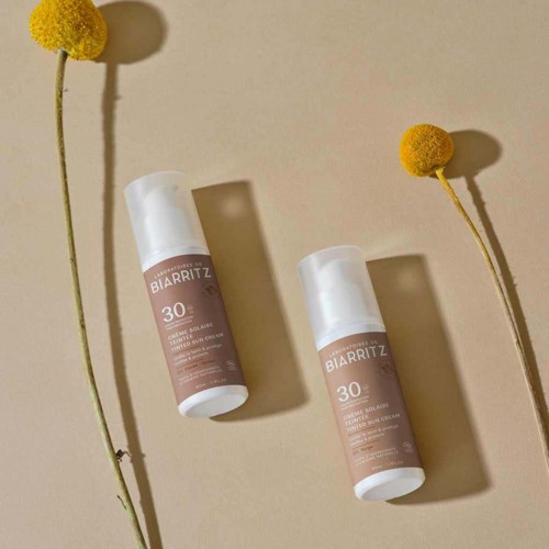 Twee zonnebrandflessen op een beige oppervlak met gele bloemen die schaduw werpen.