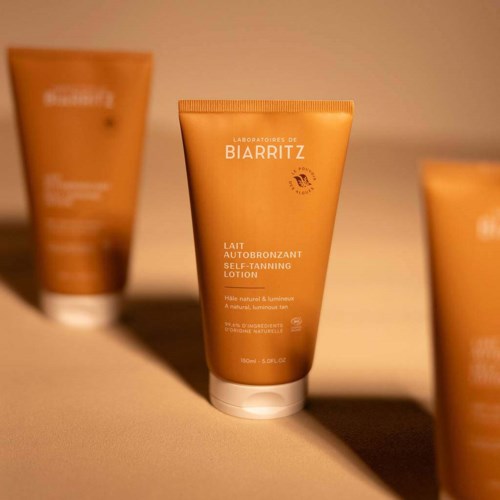 Oranje tube met witte tekst, gelabeld als zelfbruiningslotion van Biarritz, met zonthema. Twee soortgelijke tubes op de achtergrond.
