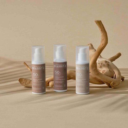 Drie flessen huidverzorging met SPF 50 op een beige oppervlak, met een gedraaide tak en houten stukjes op de achtergrond.