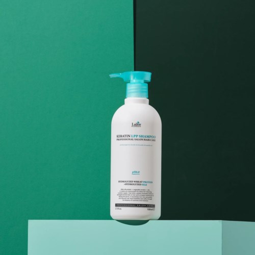 A white bottle with a pump, labeled keratin shampoo, stands on a flat surface against a green background.
