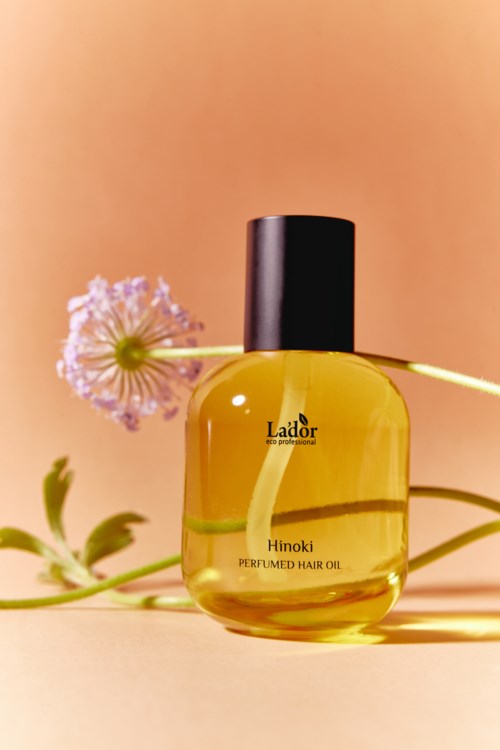 A yellow bottle of hair oil labeled 'Hinoki Perfumed Hair Oil' with a pink flower and green plant in the background.