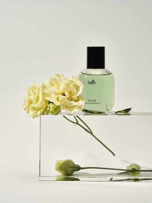 A perfume bottle with green liquid, a yellow flower, and two buds on a simple white background.
