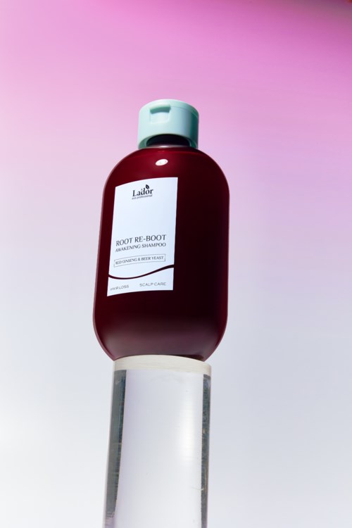Een fles shampoo genaamd 'ROOT RE-BOOT' op een witte standaard met roze en paarse achtergrond.