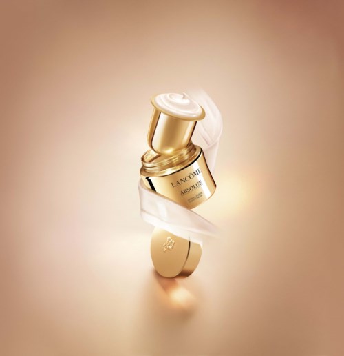 Een gouden lippenstift met een witte lint eromheen, op een zachte beige achtergrond.