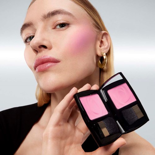 Woman with pink makeup holding a compact powder mirror, showing her face and jewelry.