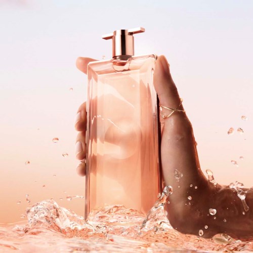 Een hand houdt een roze parfumfles met water spat eromheen, tegen een zachte pastelachtergrond.