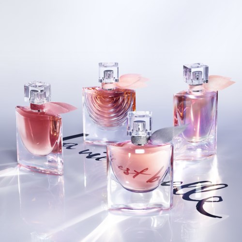 Four glass perfume bottles with pink and lavender caps are arranged on a reflective surface. The bottles are elegant and clear.