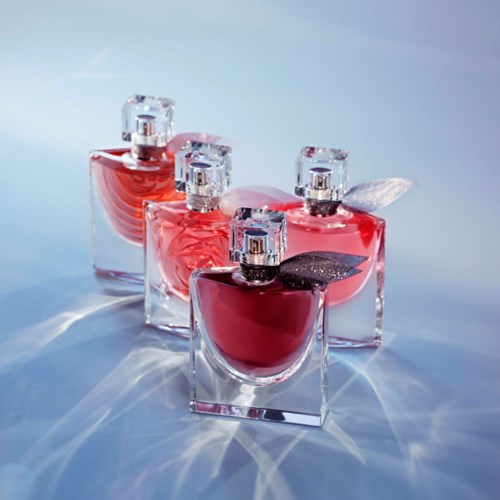 Vier glazen parfumflesjes met roze en rode vloeistoffen staan op een lichtblauwe ondergrond, reflecteren licht en werpen schaduwen.