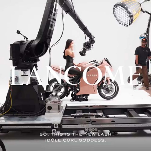 A woman wears a dress on a pink motorcycle while a robot arm films her, with bright lights and crew nearby.