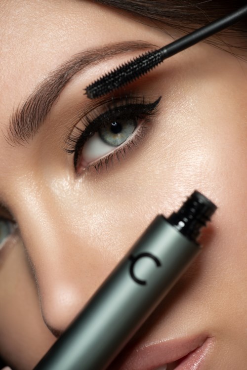 Eine Frau trägt Mascara auf ihr Auge auf, mit der Mascara-Bürste nah an den Wimpern und einem Teil des Mascara-Tubus sichtbar.