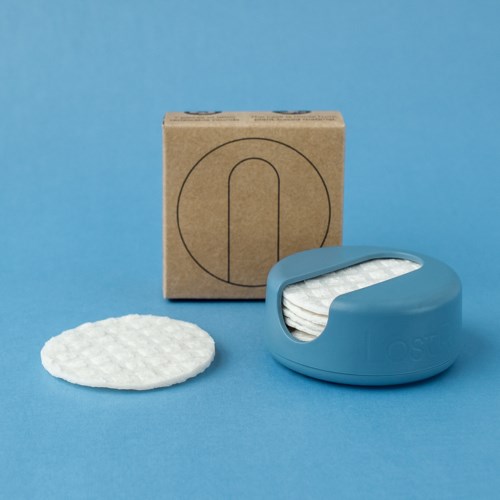 Ein blauer Behälter enthält Wattepads, mit einem Pad draußen und einer Pappschachtel dahinter auf blauem Hintergrund.