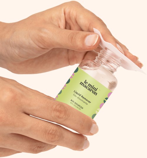 Hands hold a small transparent bottle with a green label, opened, labeled "le mini macaroon" and "liquid remover."