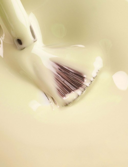A paintbrush submerged in milky white liquid, with some liquid dripping from the bristles.