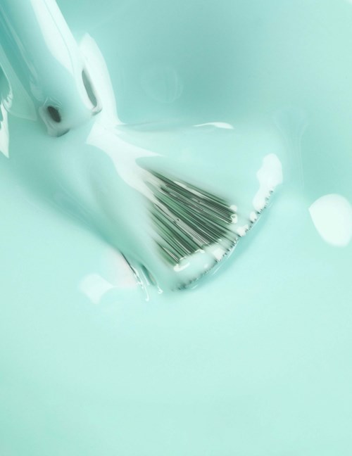 A nail polish brush is dipped in light turquoise nail polish, with some polish falling back into the bottle.