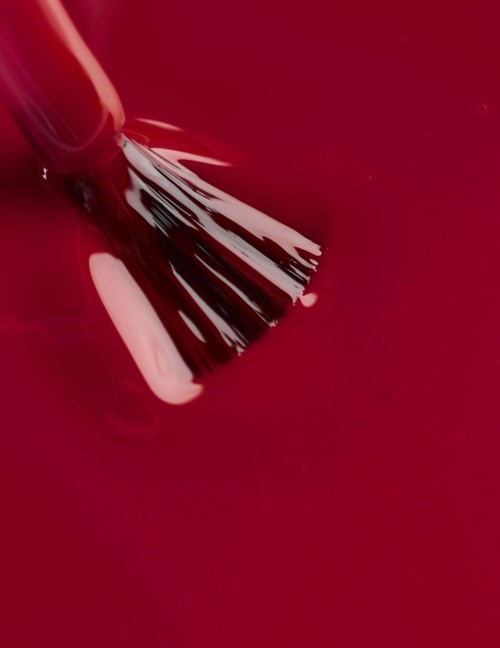 A nail polish brush is applying red nail polish with a shiny finish onto a nail.