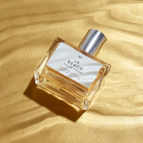 Hajuvesipullo makaa kultaista kangasta, varrellinen hopeakorkki ja valkoinen etiketti jossa lukee Le Beach Eau de Parfum.