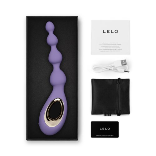 En lila, kurvad, sensorisk massager med gulddetalj, tillsammans med laddare, instruktionskort, väska och kort i boxen.