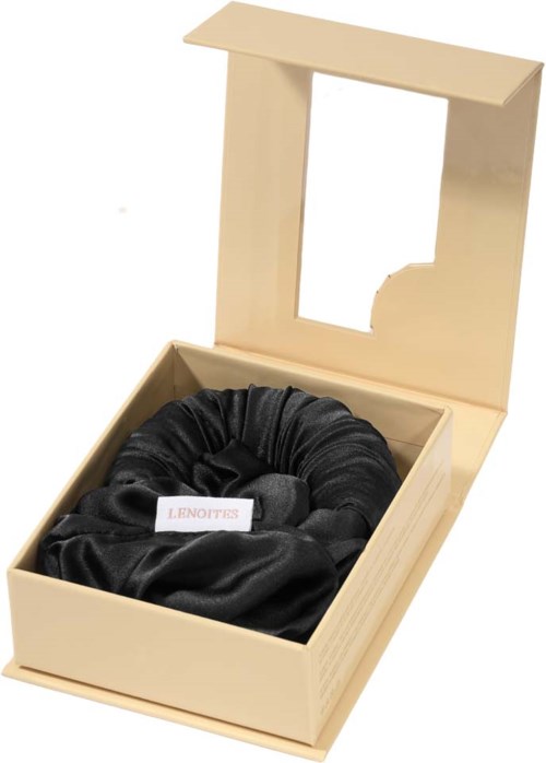 A cream-colored box with a black satin scrunchie inside, and a transparent window on the lid.