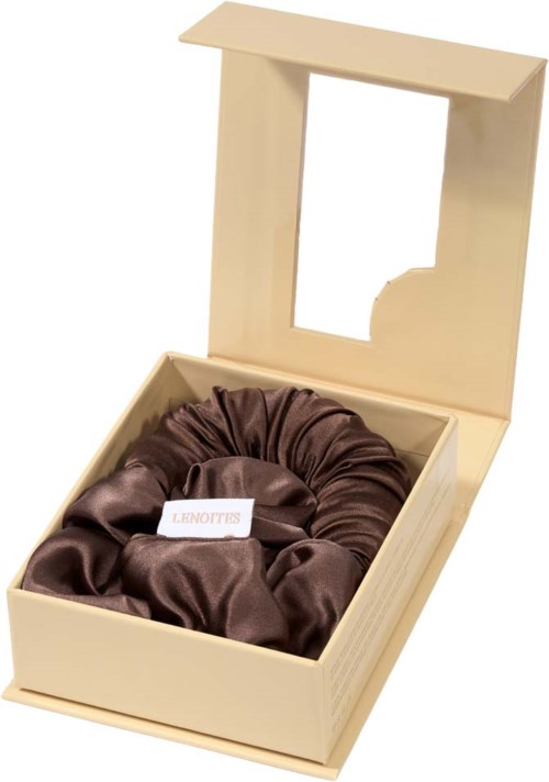 A beige box with a clear window contains a brown satin scrunchie. The box is open and looks elegant.