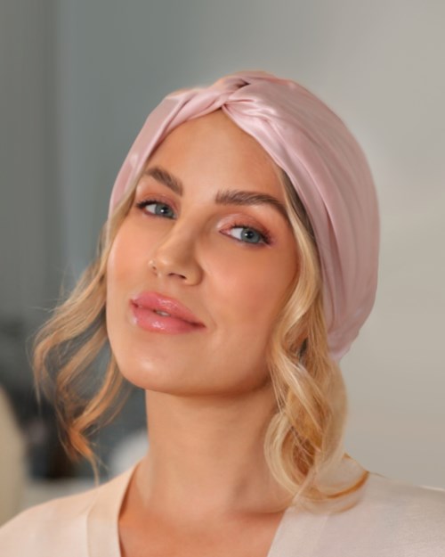 A woman with wavy blonde hair wears a pink silk headscarf and a white top.