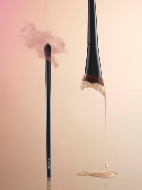 Een make-up kwast geeft poeder en vloeibone foundation af op een roze achtergrond.