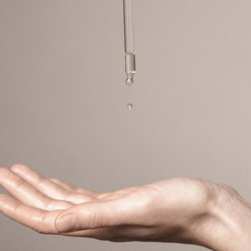 Eine Hand fängt einen Tropfen Flüssigkeit von einer Pipette ab vor einfacher Hintergrund.