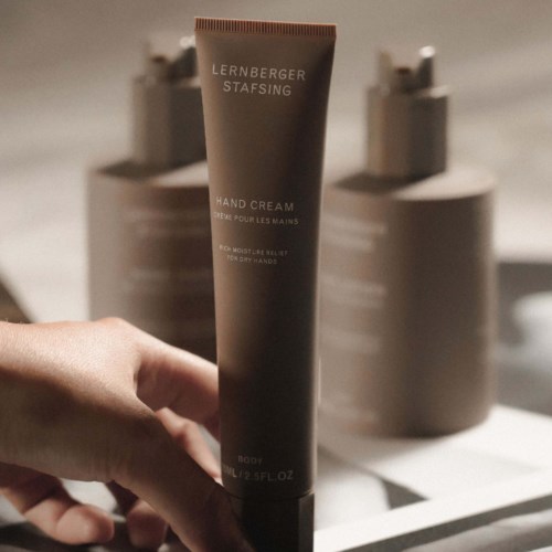 Eine Hand hält eine braune Tube mit der Aufschrift 'LERNBERGER STAFSING HAND CREAM' im Hintergrund befinden sich Flaschen.